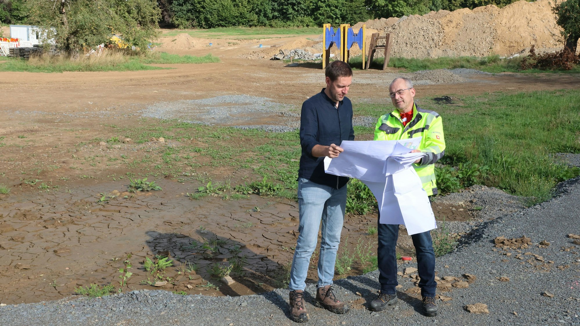 Bauamtsleiter Jan Foerster und Jörn Renke vom Planungsbüro Schumacher stehen auf einem freien Gelände und schauen auf einen Plan.