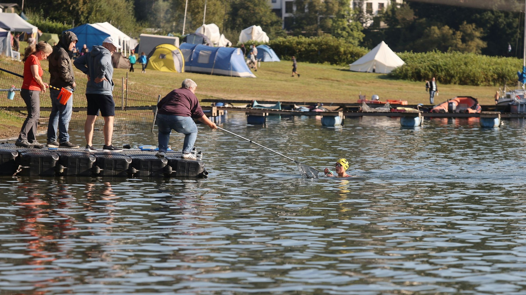 Zu sehen ist ein Mann, der einer Schwimmerin per Kescher eine Trinkflasche ins Wasser reicht.