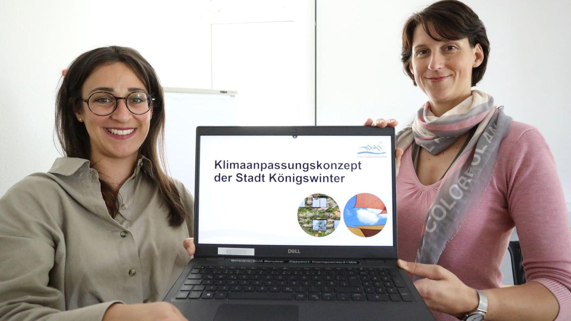 Zwei junge Frauen halten einen Laptop, auf dessen Monitor der Schriftzug „Klimaanpassungskonzept der Stadt Königswinter“ steht.