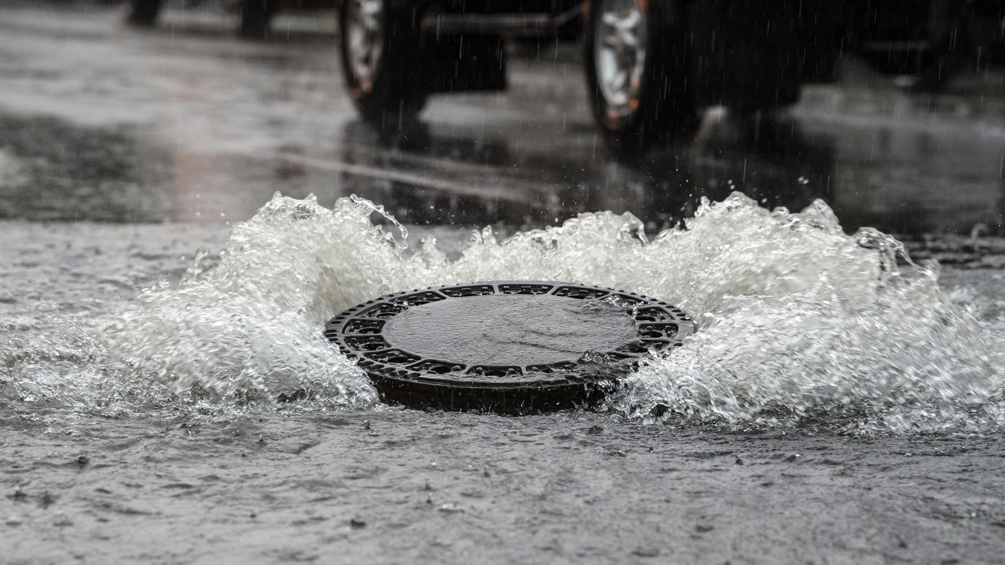 Ein Gullydeckel wird auf einer überfluteten Straße vom Wasserdruck hochgehoben. (Symbolbild)