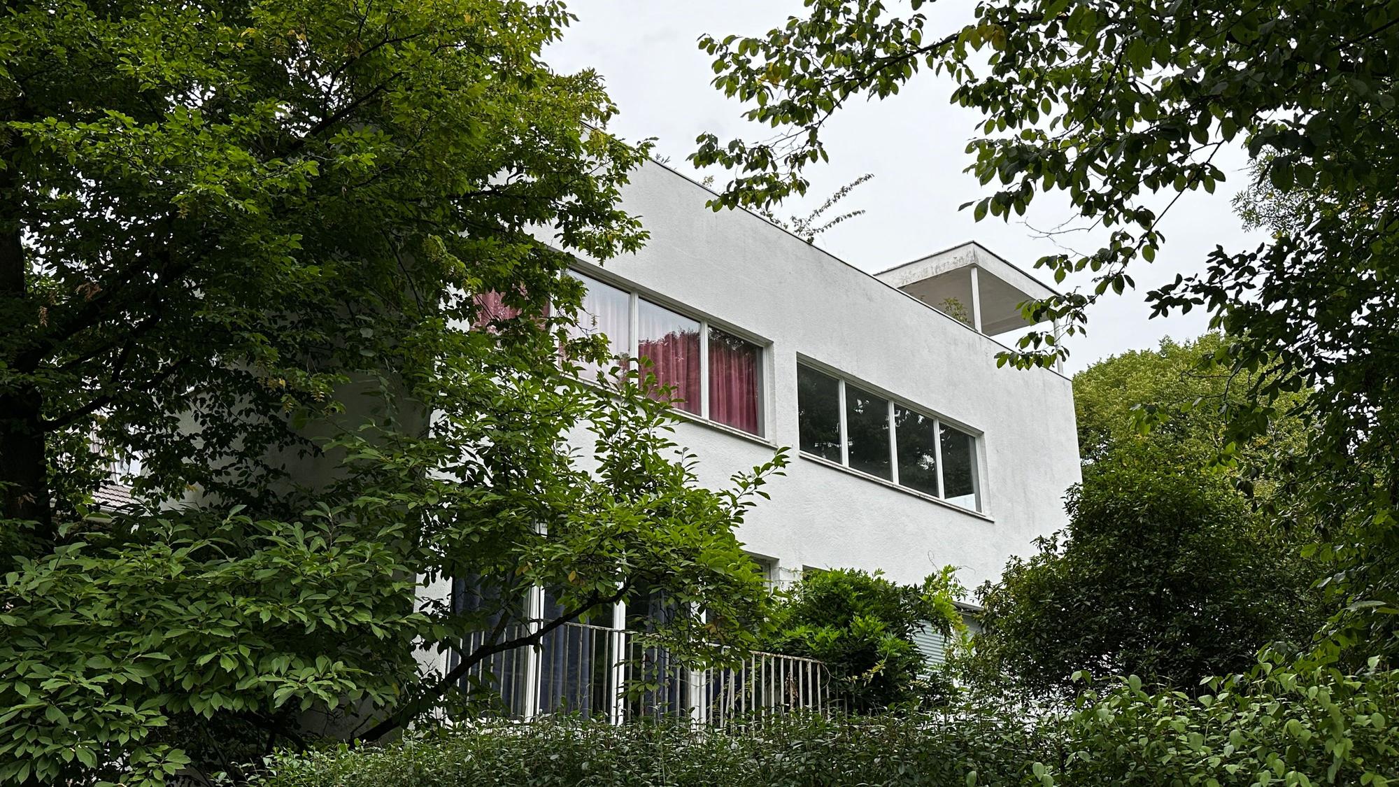 Ein weißes Gebäude im Bauhaus-Stil wird von Bäumen und Hecken eingerahmt.