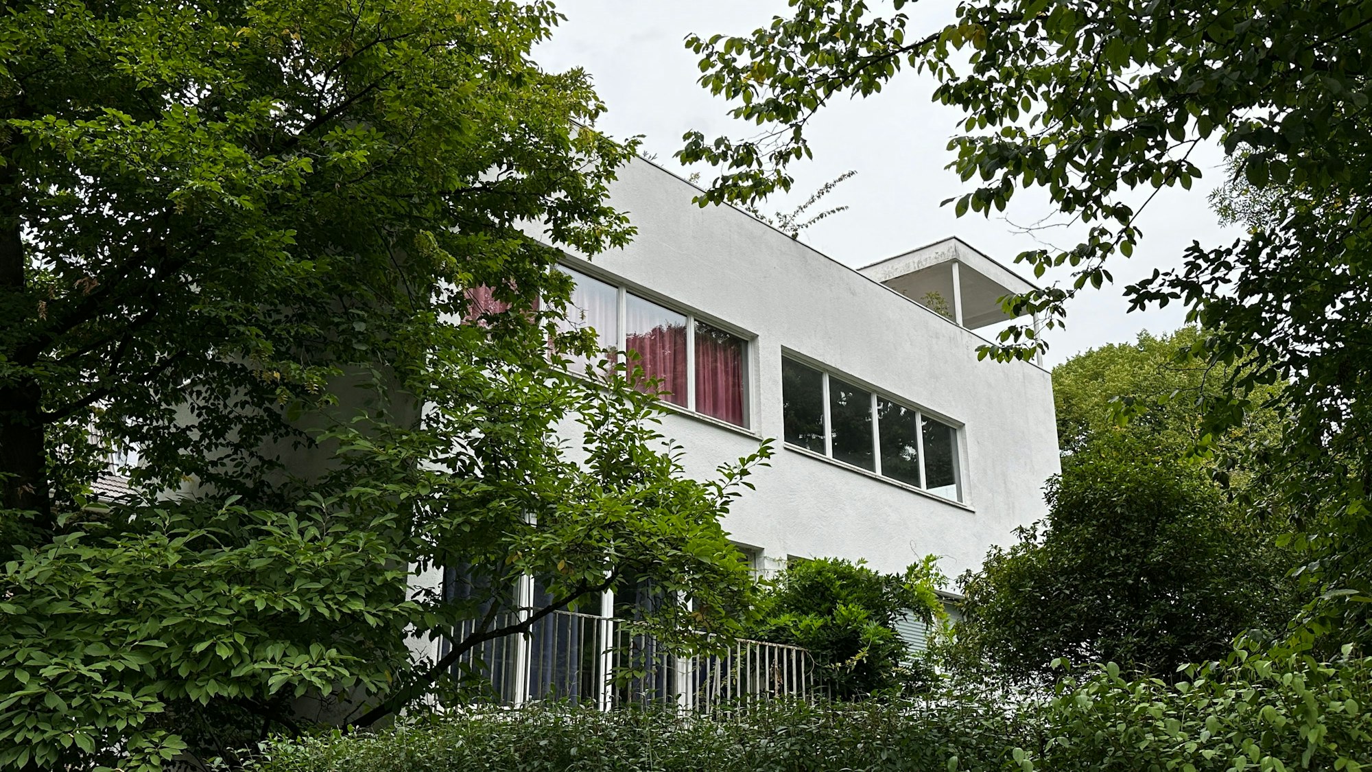 Ein weißes Gebäude im Bauhaus-Stil wird von Bäumen und Hecken eingerahmt.