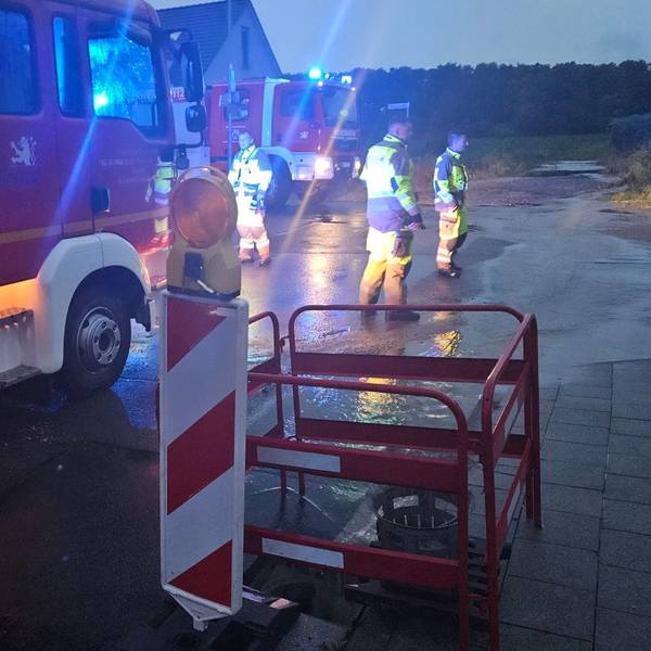 Zu sehen sind Feuerwehrleute und Feuerwehrfahrzeuge auf einer mit Wasser überfluteten Straße.