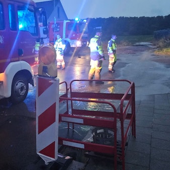 Zu sehen sind Feuerwehrleute und Feuerwehrfahrzeuge auf einer mit Wasser überfluteten Straße.