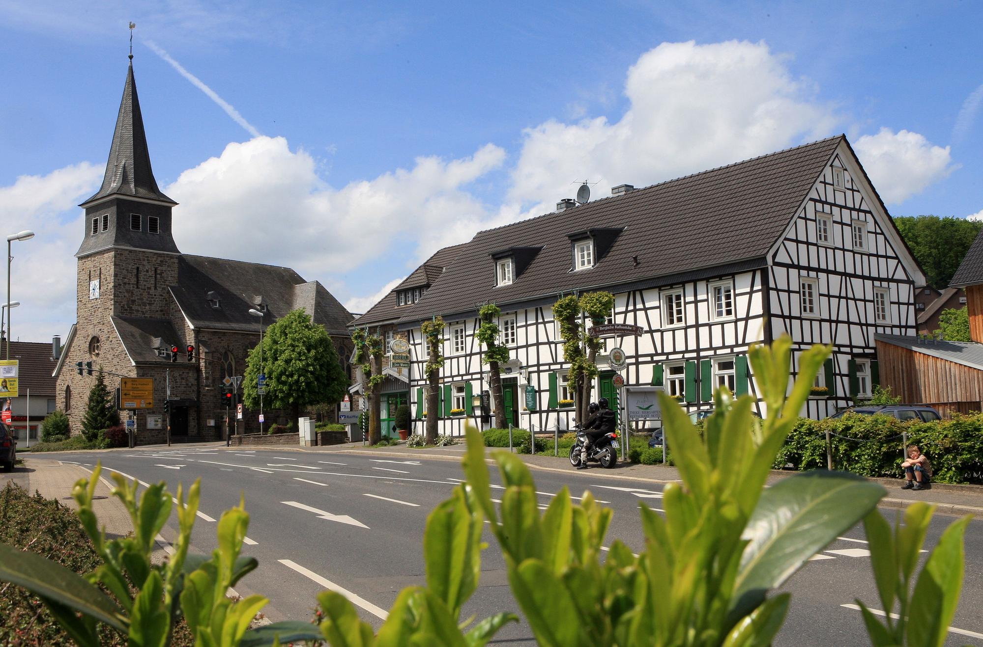 Eine Kirche mit trutzigem Turm aus Bruchstein. Daneben ein historisches Fachwerkhaus. Davor eine Straße.