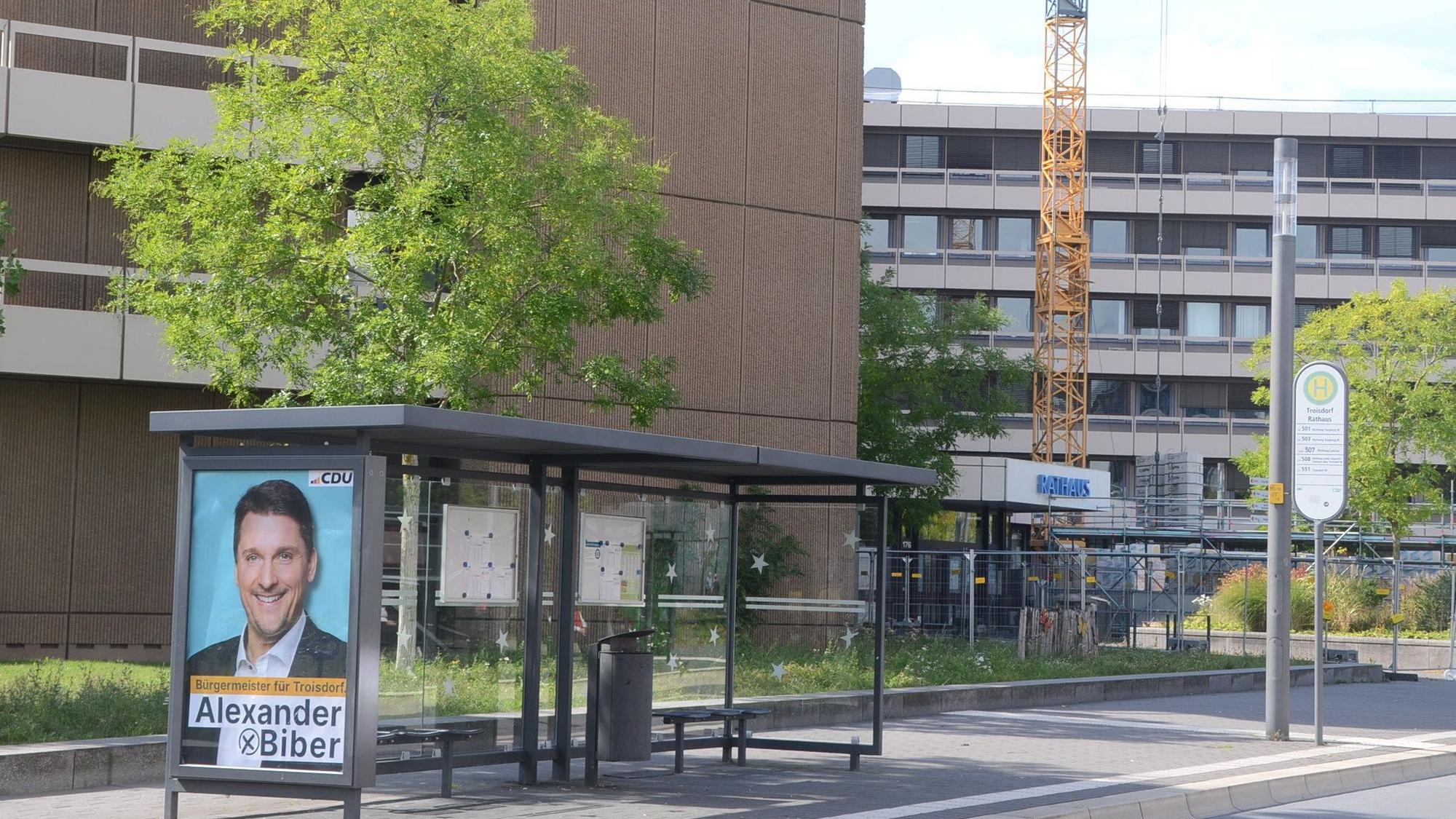 Ein Plakat mit Wahlwerbung an einer Bushaltestelle. Im Hintergrund ein Gebäude mit der Aufschrift Rathaus.