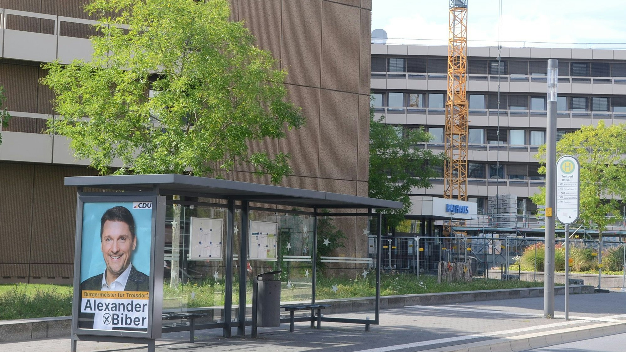 Ein Plakat mit Wahlwerbung an einer Bushaltestelle. Im Hintergrund ein Gebäude mit der Aufschrift Rathaus.