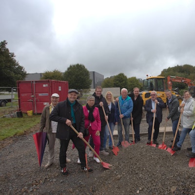 Mit roten Schaufeln stehen zwölf Personen im Halbkreis und machen symbolisch den Spatenstich.