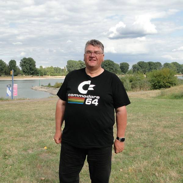 Ein Mann mit Brille und schwarzer Hose und Shirt steht auf einer Wiese am Rhein.
