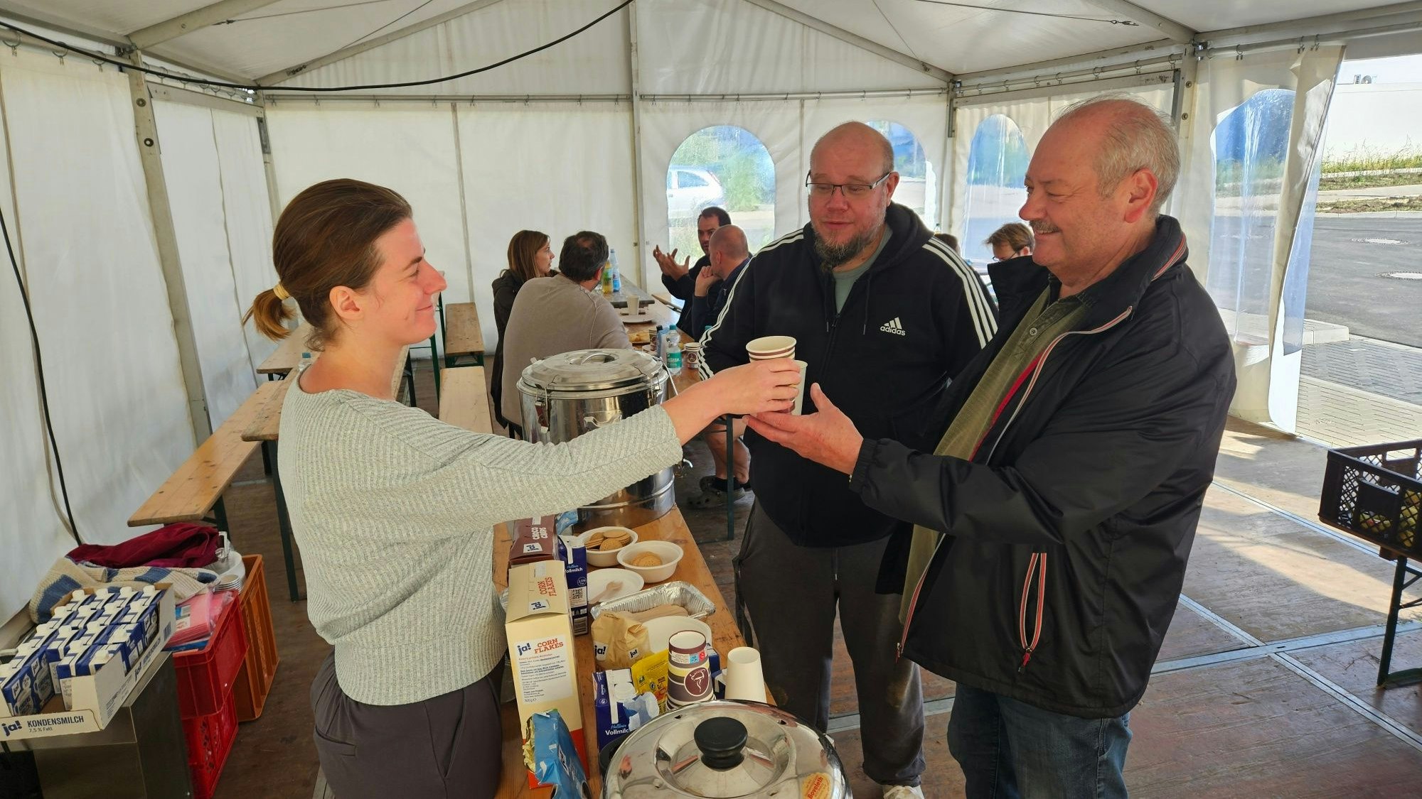 Zu sehen ist eine Frau, die in einem Zelt Kaffee an zwei Männer ausgibt.