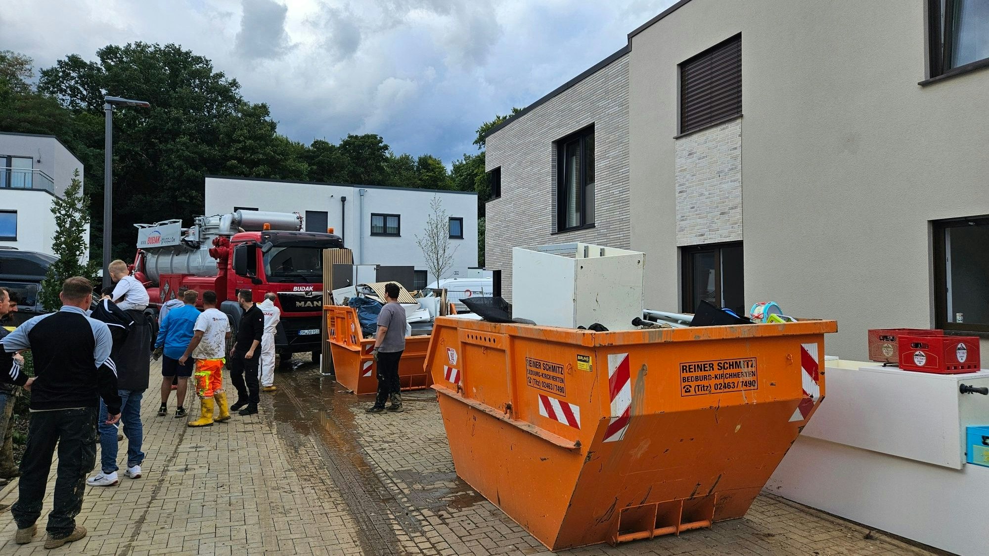 Zu sehen sind Container voller Sperrmüll in einer Naubausiedlung, Menschen stehen auf der Straße.