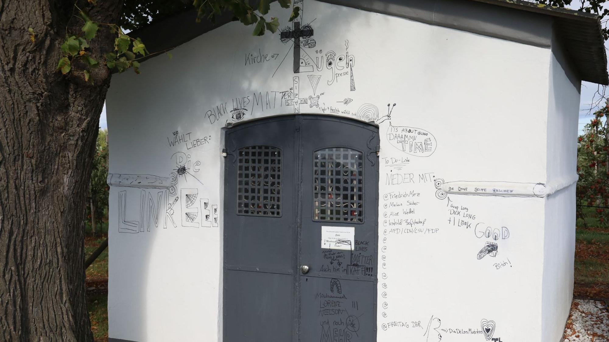 Eine kleine weiße Kapelle mit schwarzer Eisentür, über der ein Kreuz hängt.