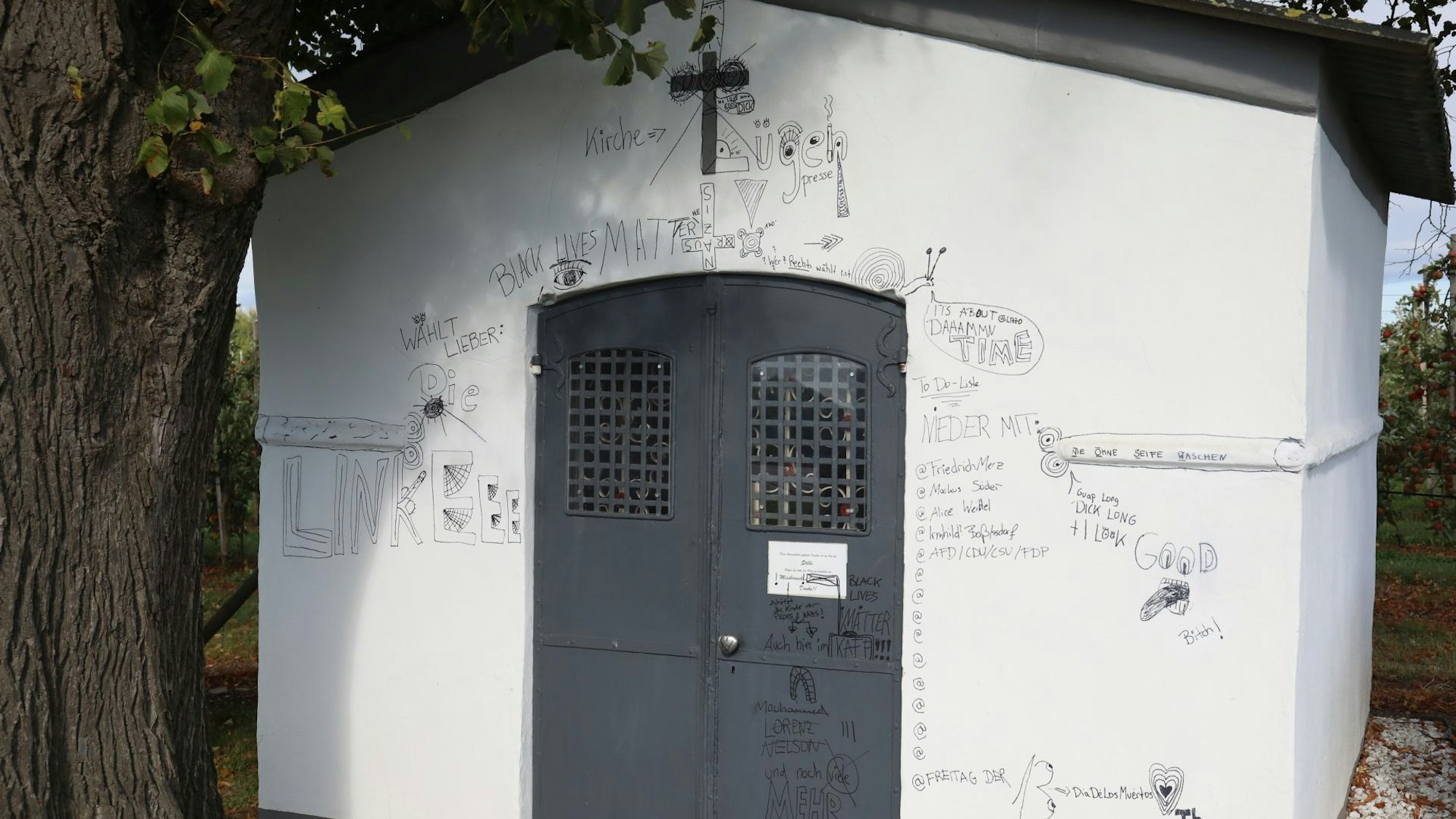 Eine kleine weiße Kapelle mit schwarzer Eisentür, über der ein Kreuz hängt.