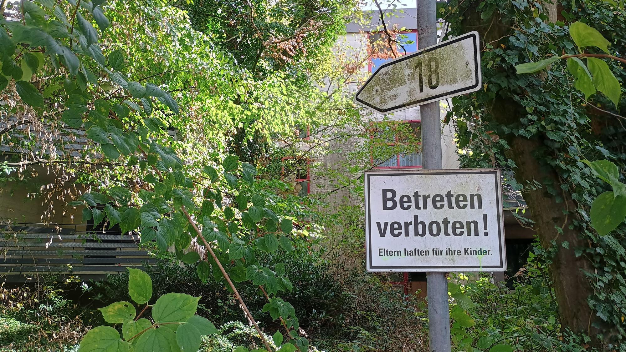 Ein Schild mit der Nummer 18, darunter ein Schild mit der Aufschrift: „Betreten verboten! Eltern haften für ihre Kinder“. Im Hintergrund das Gebäude, stark von Pflanzen verdeckt.