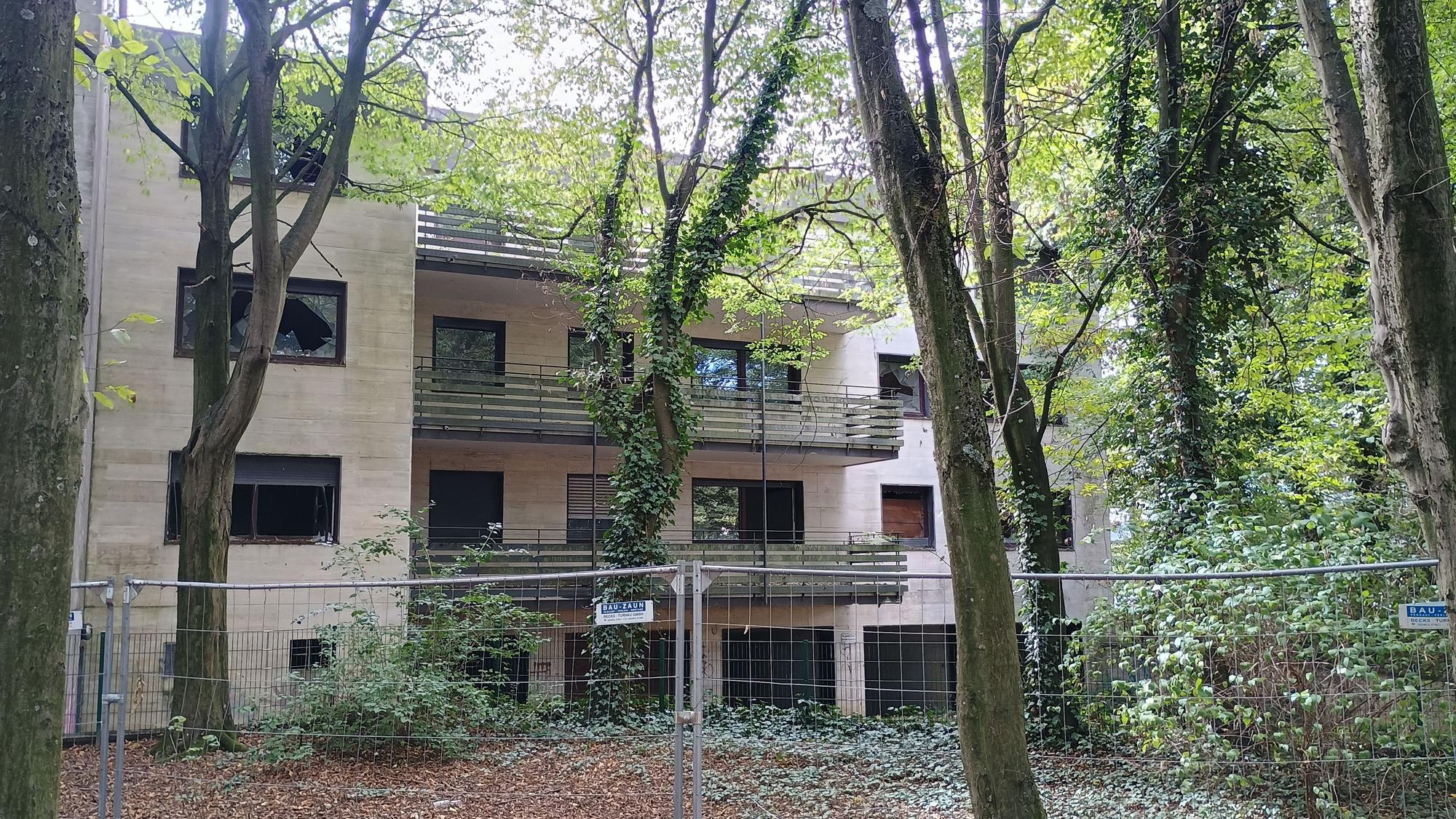 Das Bild zeigt das umzäunte Gebäude des Wohnparks. Viele Fenster sind eingeschlagen, einige Bäume verdecken die Sicht auf das Haus.