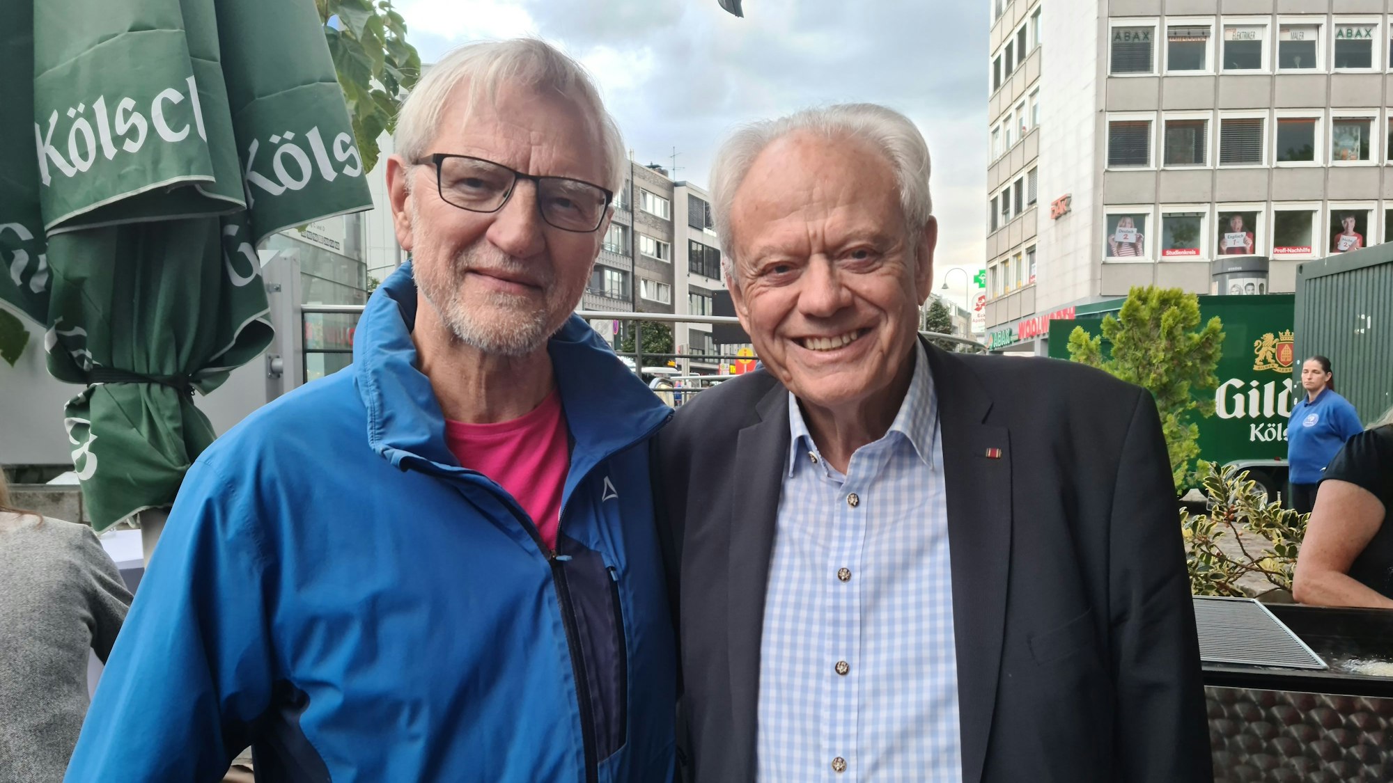Ein Mann im blauen Anorak steht neben einem älteren Mann in hellblau-kariertem Hemd und dunkelblauem Anzug.