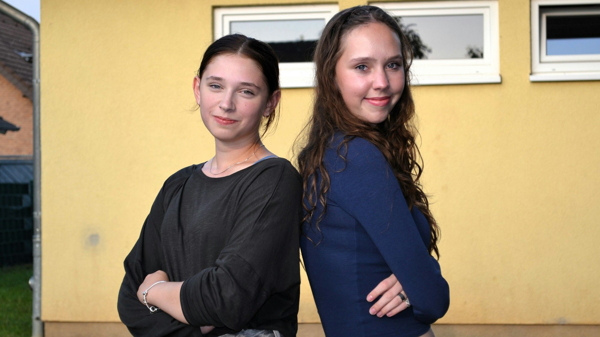 Zwei junge Mädchen stehen Rücken an Rücken und schauen in die Kamera.