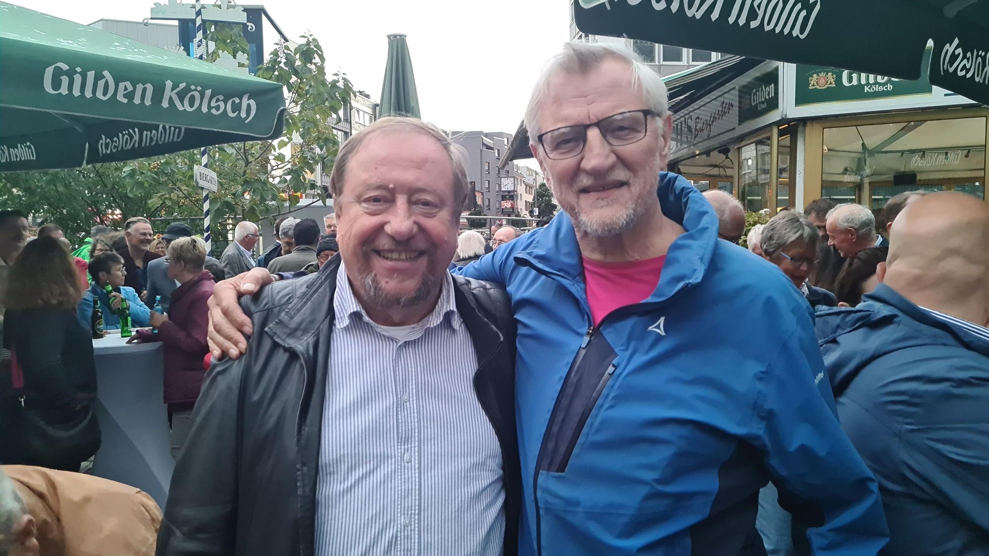 Ein Mann im hellblau-weiß gestreiftem Hemd und schwarzer Jacke steht neben einem Mann mit Brille im blauen Anorak.