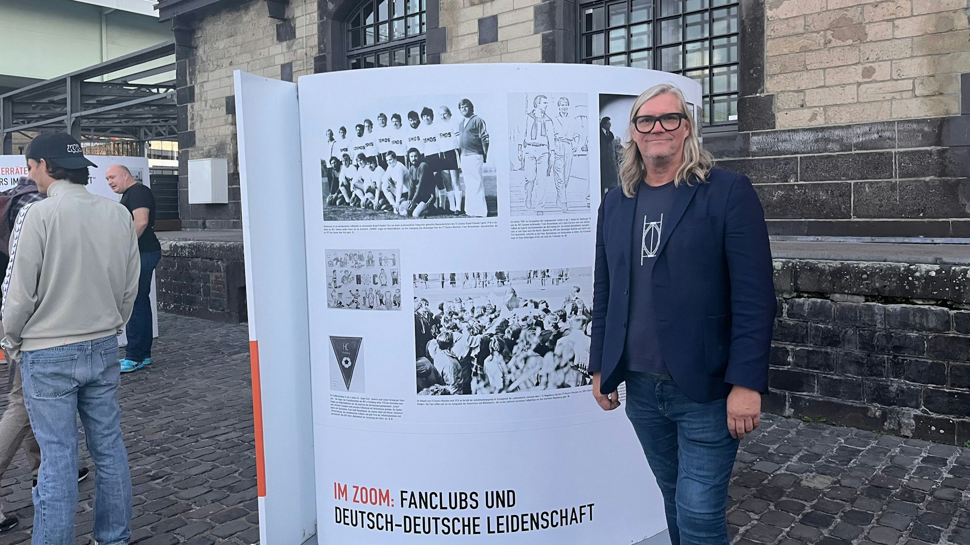 Ein Mann steh vor einer Infotafel mit schwarz-weiß-Fotografien von Fußballfans mit Kosmos-Aufschrift.