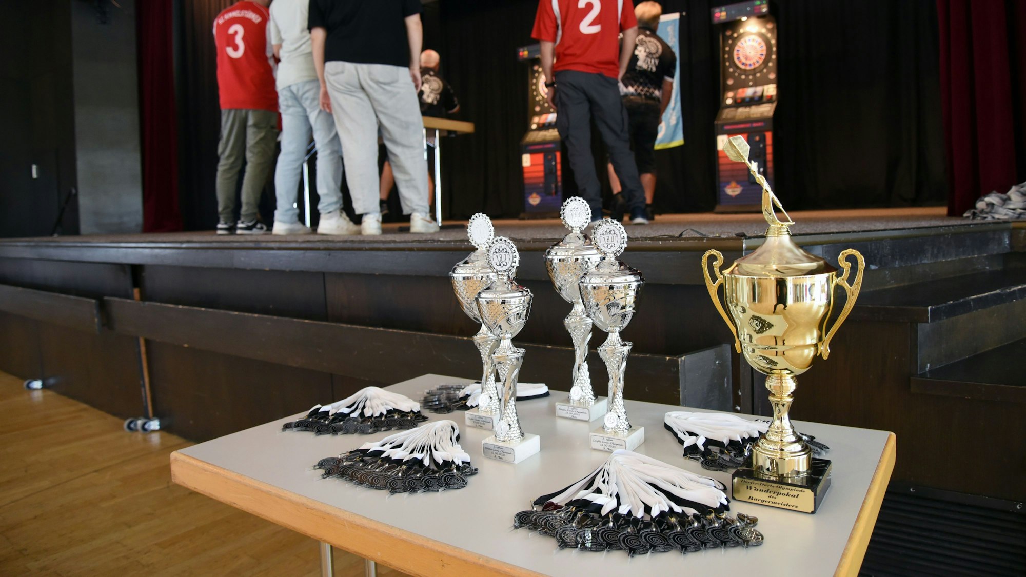 Pokale und Medaillen in Dartpfeil-Form stehen und liegen auf einem Tisch.