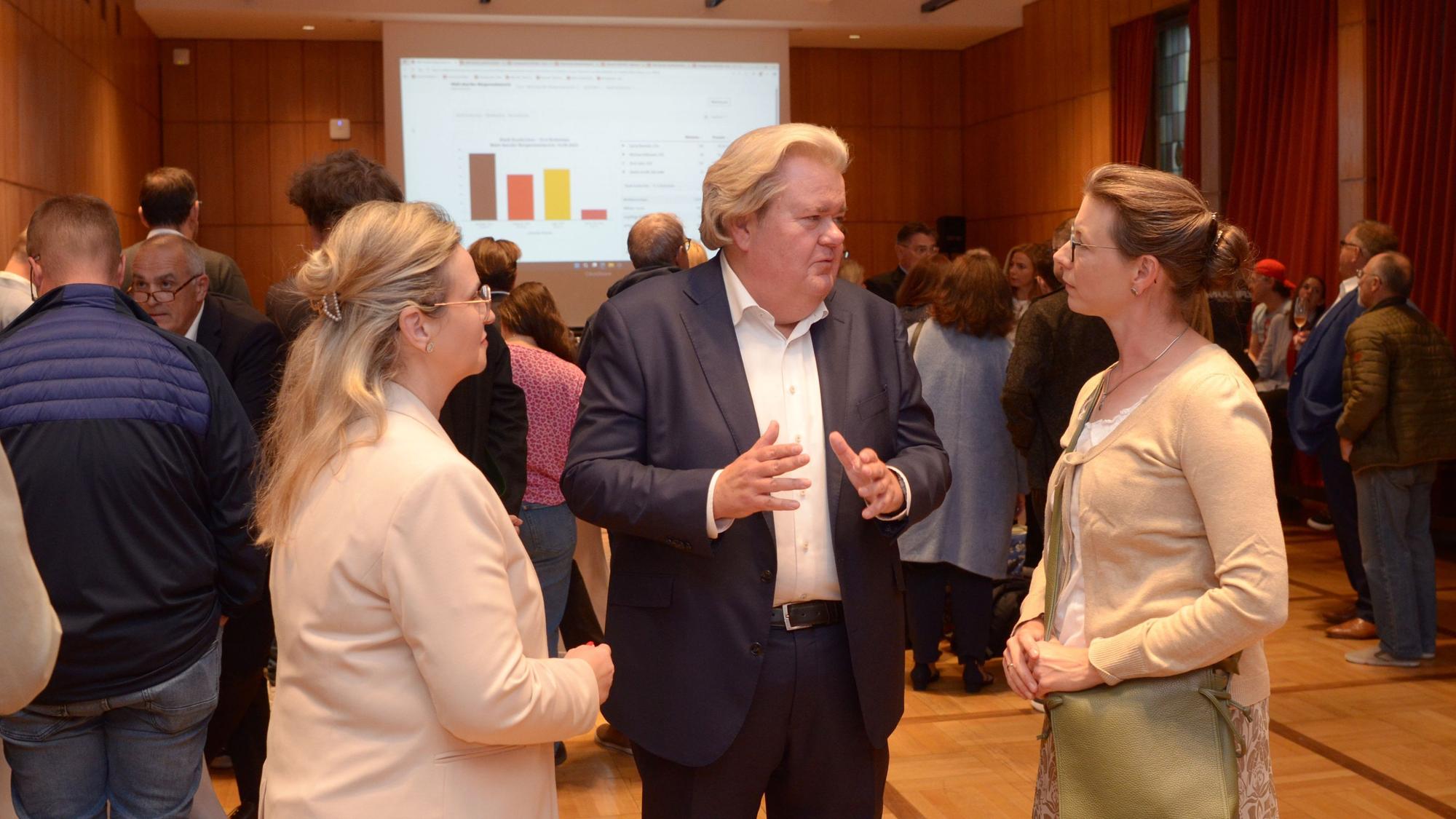 Zwei Frauen und ein Mann unterhalten sich miteinander. Im Hintergrund stehen weitere Menschen, einige schauen auf eine Leinwand mit Wahlergebnissen.