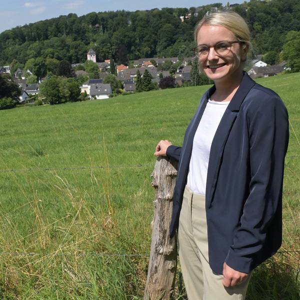 Eine junge Frau mit blonden, glatten Haaren und Brille steht vor einer Wiese mit Blick auf das historische Zentrum von Odenthal.