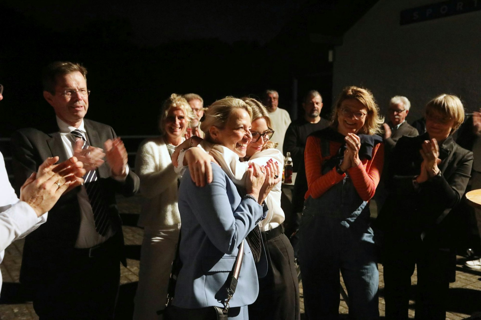 Zwei Frauen umarmen sich, mehrere Menschen applaudieren.