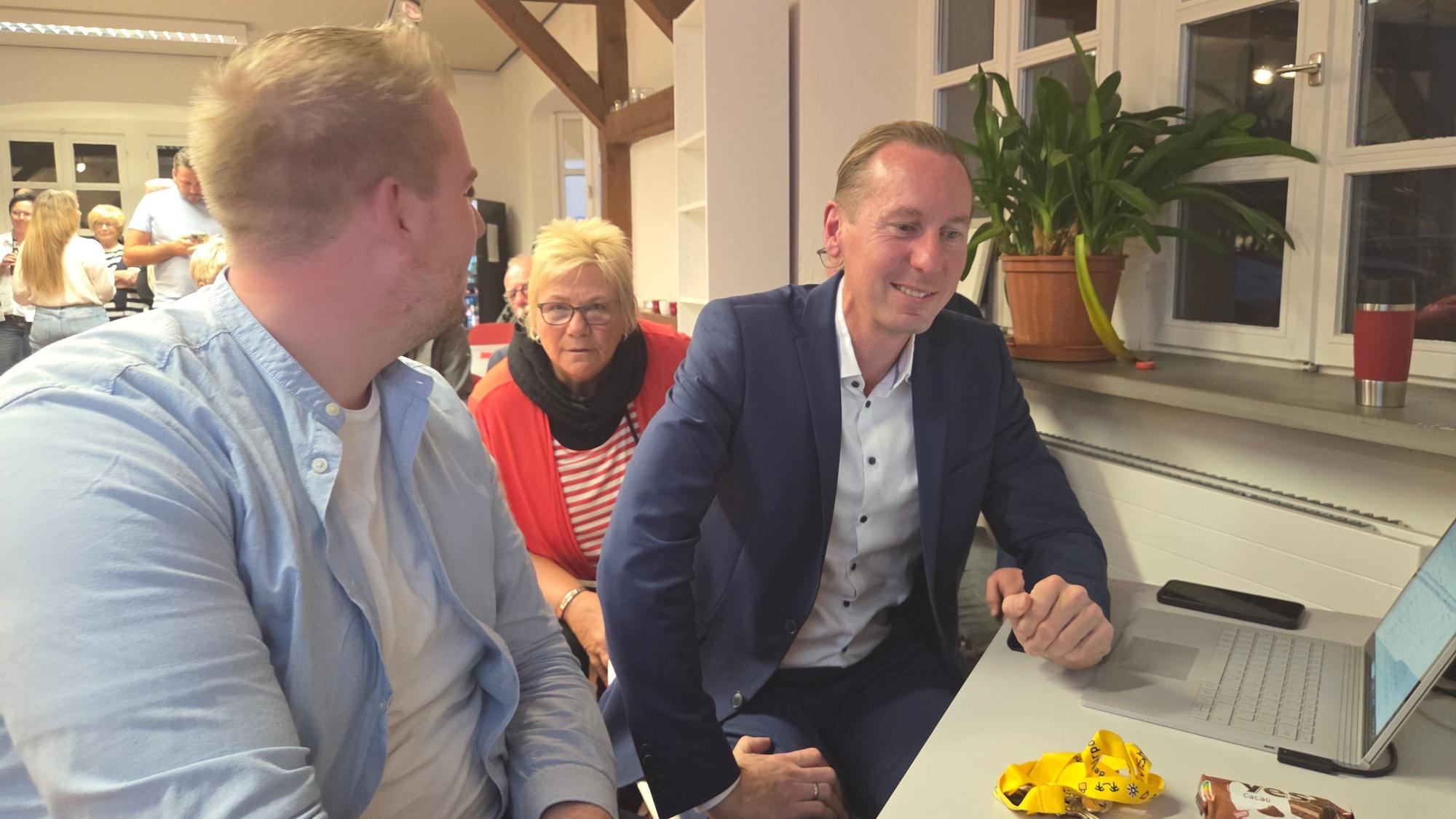 Zwei Männer und eine Frau an einem Schreibtisch mit einem aufgeklappten Laptop.