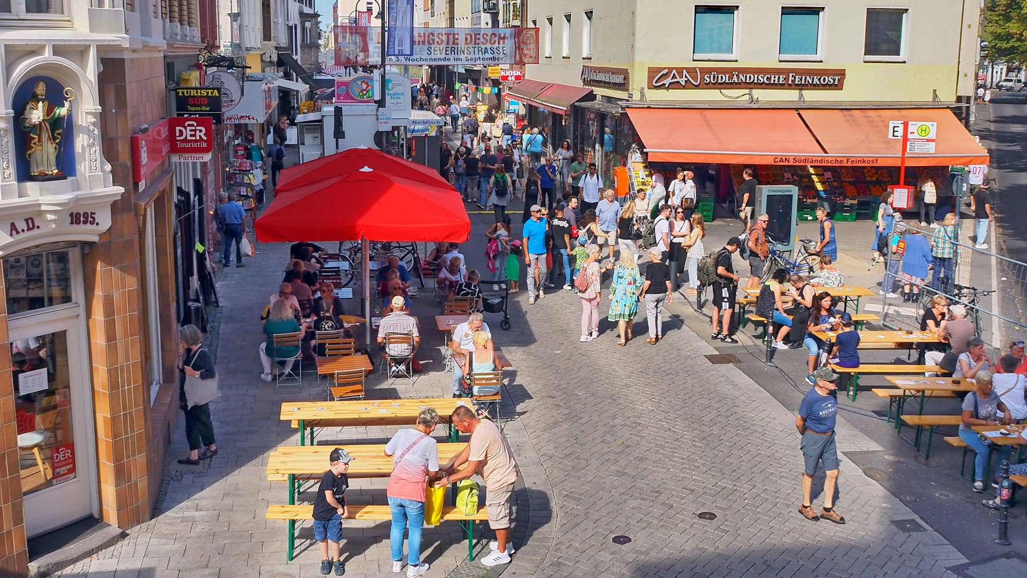 Links und rechst von einer schmalen Straße stehen Bierbänke, Menschen laufen die Straße entlang oder sitzen an den Tischen.