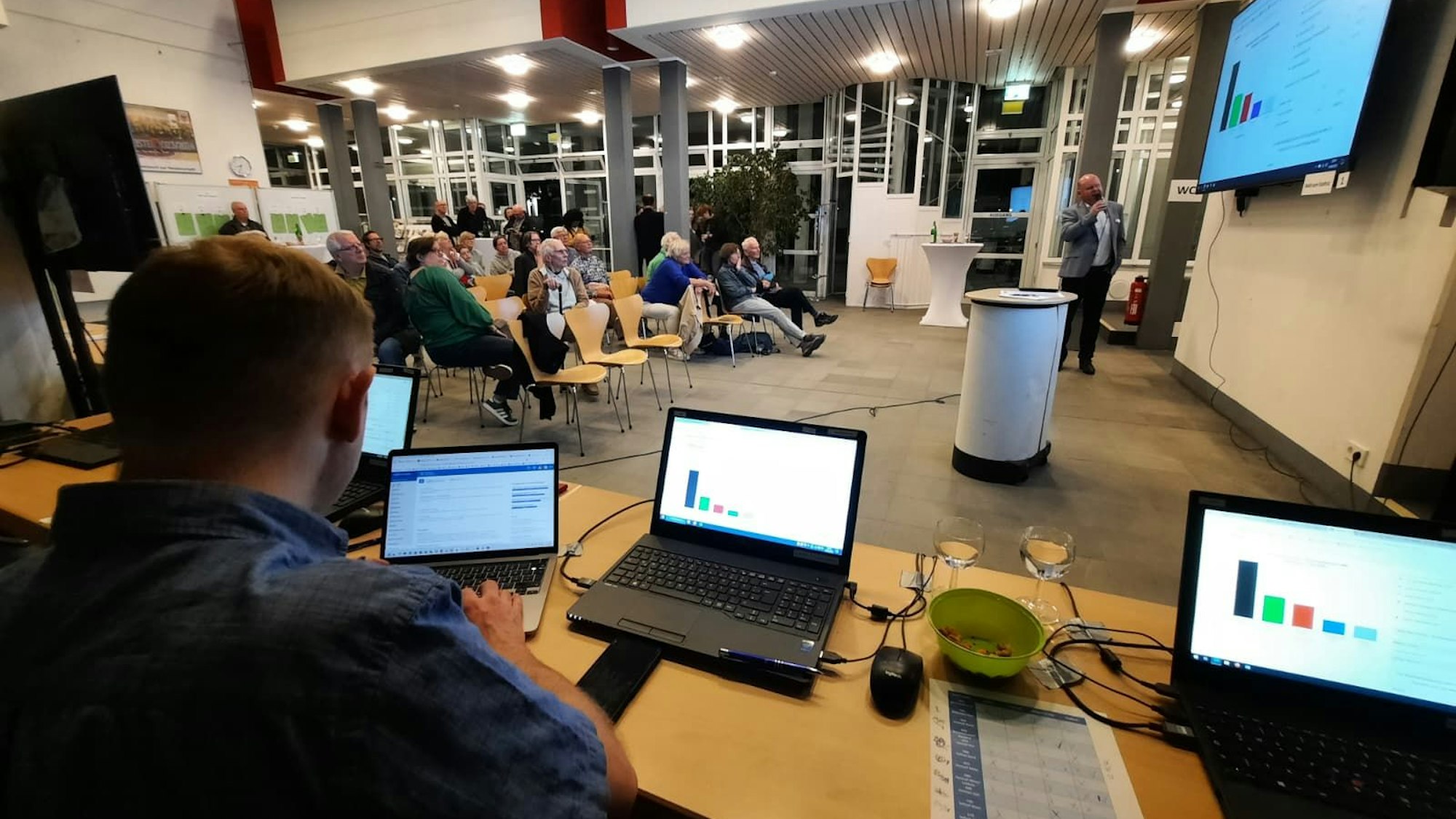 Ein Tisch mit mehreren Computern im Vordergrund, ein großer Bildschirm an der Wand, davor sitzen Menschen auf Stühlen.