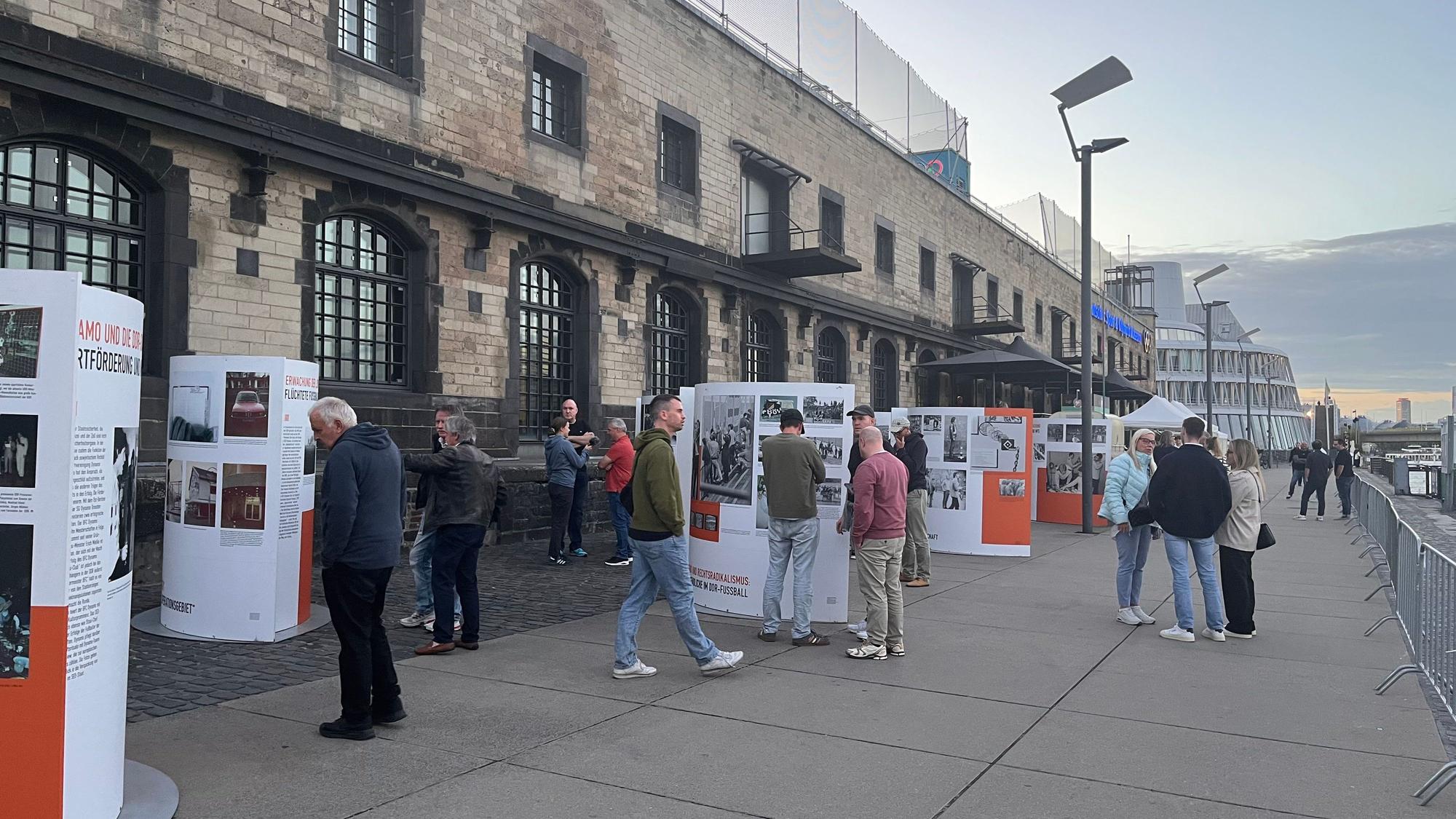 Auf der Promenadenstraße am Rhein schauen sich mehrere Menschen vor dem Museumsgebäude Infotafeln mit schwarz-weiß-Bildern an.