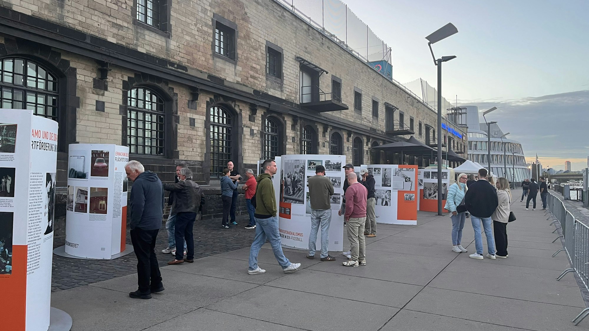 Auf der Promenadenstraße am Rhein schauen sich mehrere Menschen vor dem Museumsgebäude Infotafeln mit schwarz-weiß-Bildern an.