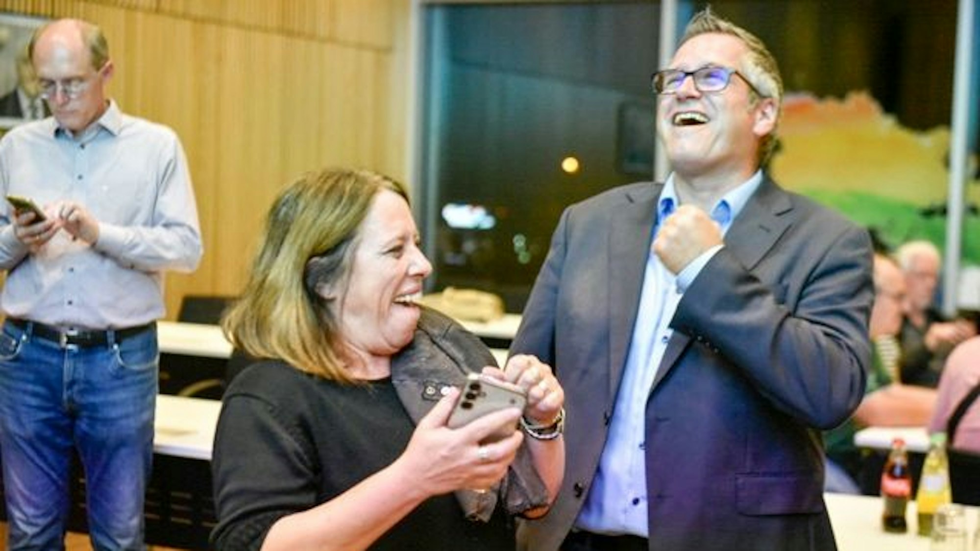 Zu sehen sind ein Mann und eine Frau, die feiern, im Hintergrund tippt ein Mann auf seinem Handy.