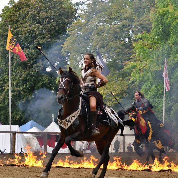 Die kostümierte Patricia Beissel reitet auf einem Pferd. Im Hintergrund ein Reiter, der auf seinem Pferd durch Feuer läuft.