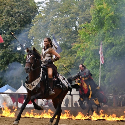 Die kostümierte Patricia Beissel reitet auf einem Pferd. Im Hintergrund ein Reiter, der auf seinem Pferd durch Feuer läuft.