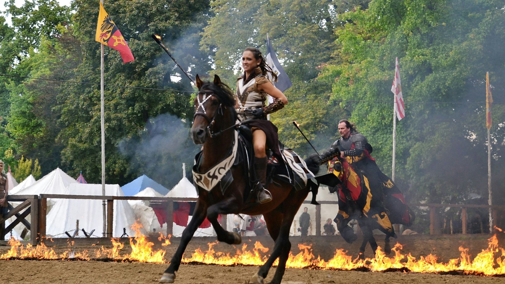 Die kostümierte Patricia Beissel reitet auf einem Pferd. Im Hintergrund ein Reiter, der auf seinem Pferd durch Feuer läuft.
