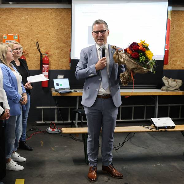 Norbert Crump hat einen Blumenstrauß in der Hand und spricht in ein Mikrofon. Links neben ihn stehen mehrere Verwaltungsmitarbeiterinnen.