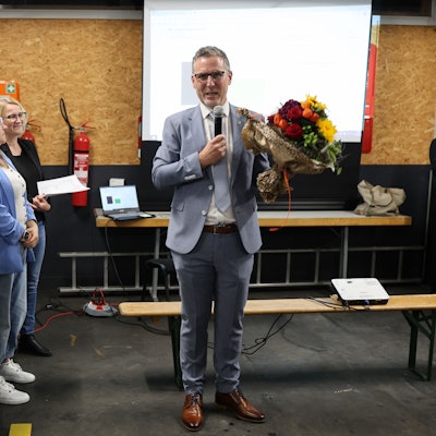 Norbert Crump hat einen Blumenstrauß in der Hand und spricht in ein Mikrofon. Links neben ihn stehen mehrere Verwaltungsmitarbeiterinnen.