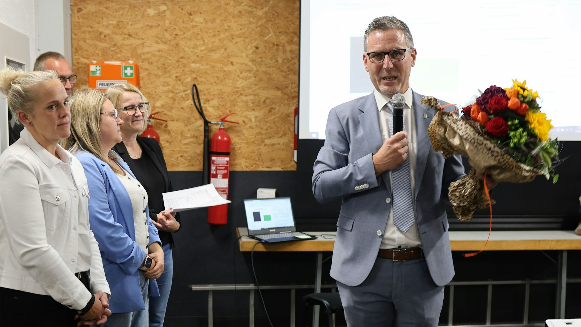 Norbert Crump hat einen Blumenstrauß in der Hand und spricht in ein Mikrofon. Links neben ihn stehen mehrere Verwaltungsmitarbeiterinnen.