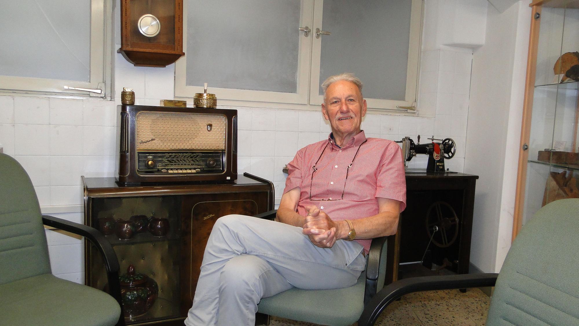 Ein älterer Herr mit grauem Haar und Oberlippenbart sitzt auf einem altmodischen Sessel, neben ihm auf einer Kommode steht ein antikes Radio.
