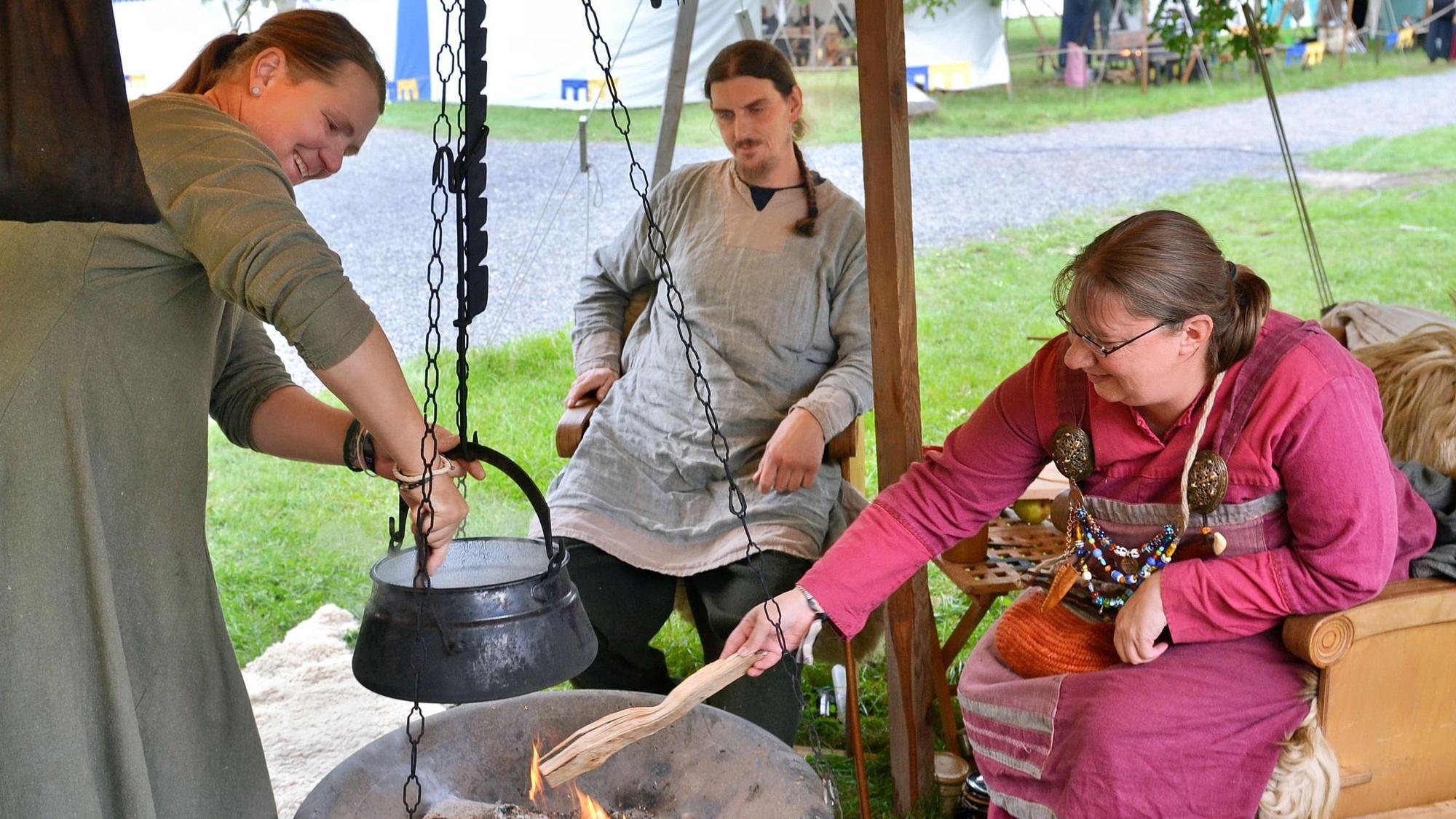 Eine Gruppe sitzt um einen Schwenkgrill herum. Eine Frau rührt in einem Topf.