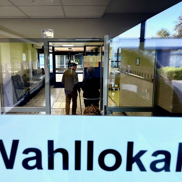 Der Blick geht in einen gefliesten Flur, in dem ein Senior mit einem Stock steht. Auf einer Tür ist der Schriftzug „Wahllokal“.