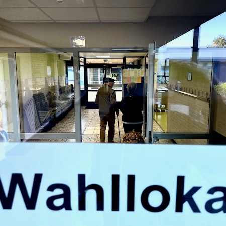 Der Blick geht in einen gefliesten Flur, in dem ein Senior mit einem Stock steht. Auf einer Tür ist der Schriftzug „Wahllokal“.