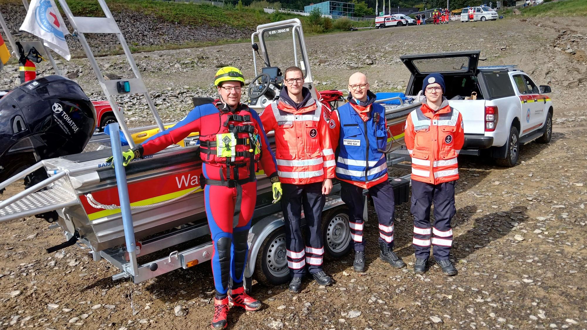 Vier Helfer des DRK, drei davon in normaler Einsatzkleidung, einer im Taucheranzug, stehen an einem Rettungsboot, das auf einem Anhänger hinter einem Einsatzfahrzeug liegt.