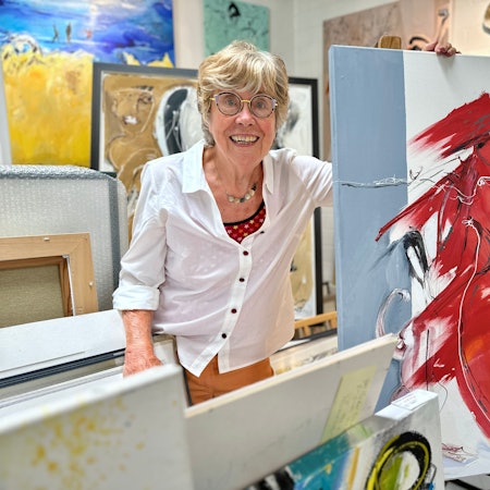 Eine Frau mit Brille und weißer Bluse steht zwischen vielen farbenfrohen Bildern.