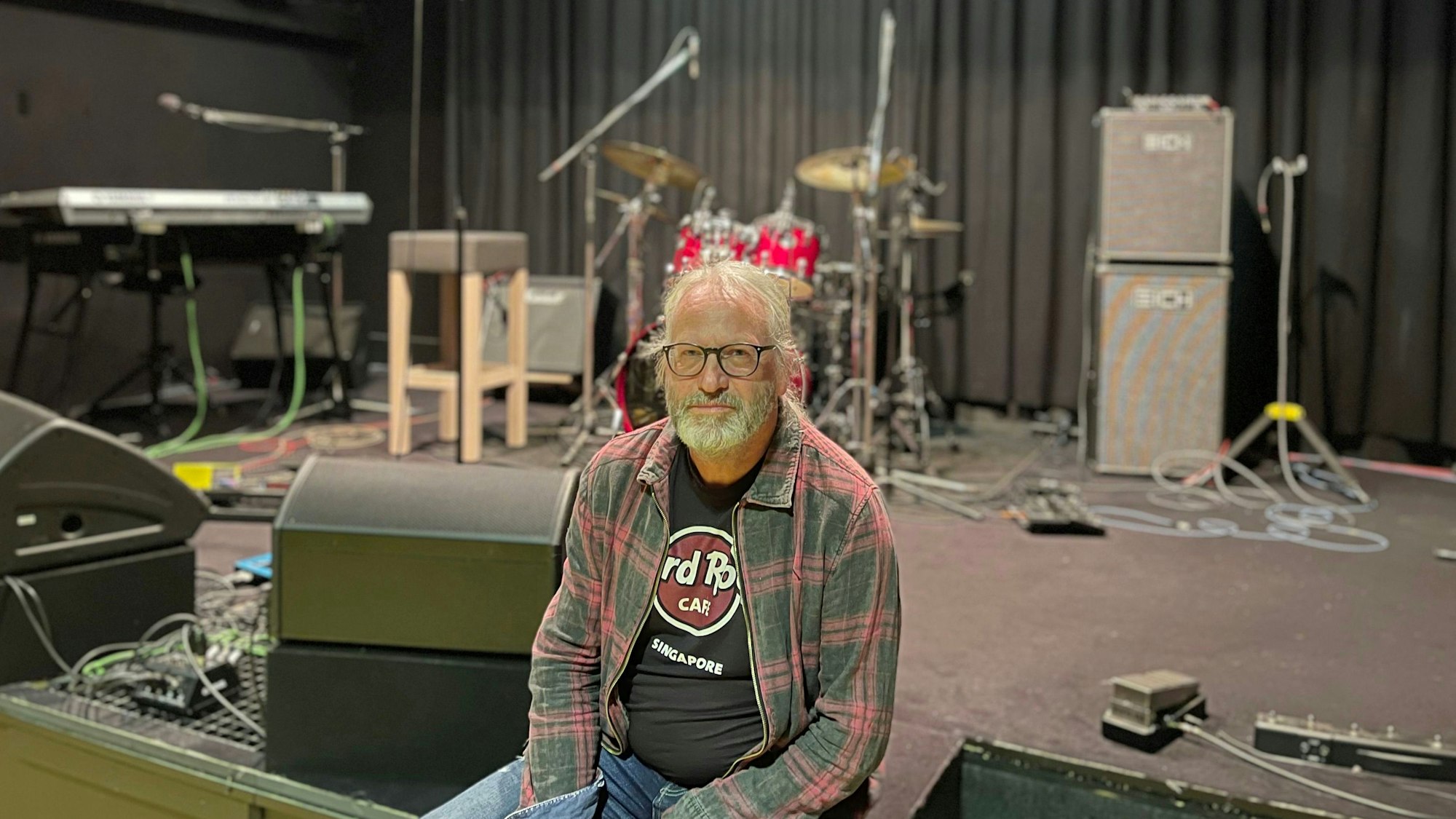 Lehser sitzt am Bühnenrand, hinter ihm ein Schlagzeug, Boxen und anderes Equipment.