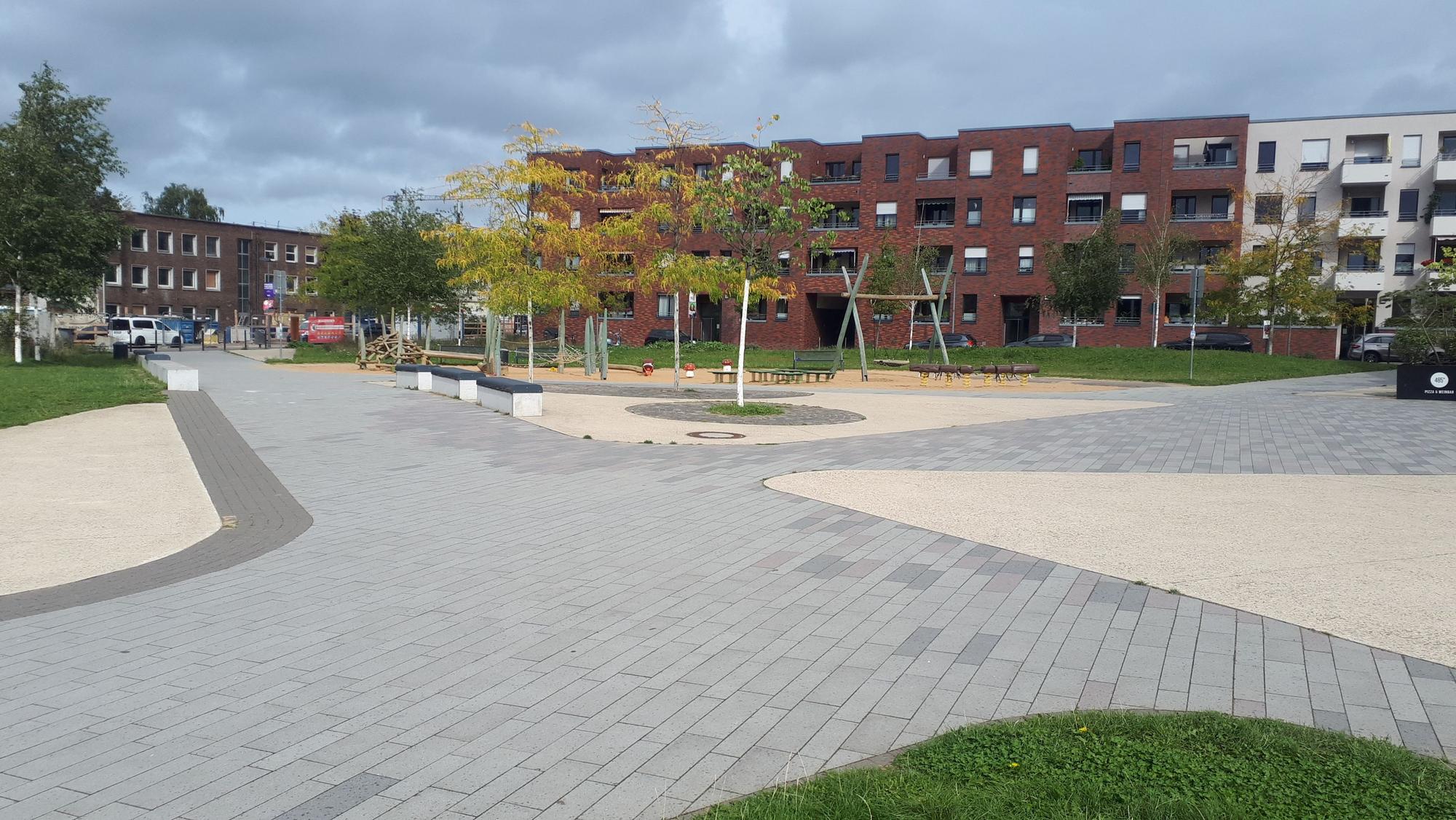 Ein gepflasterter Platz mit ein paar kleinen Bäumen und einem Spielplatz. Im Hintergrund sind Wohnhäuser zu sehen.