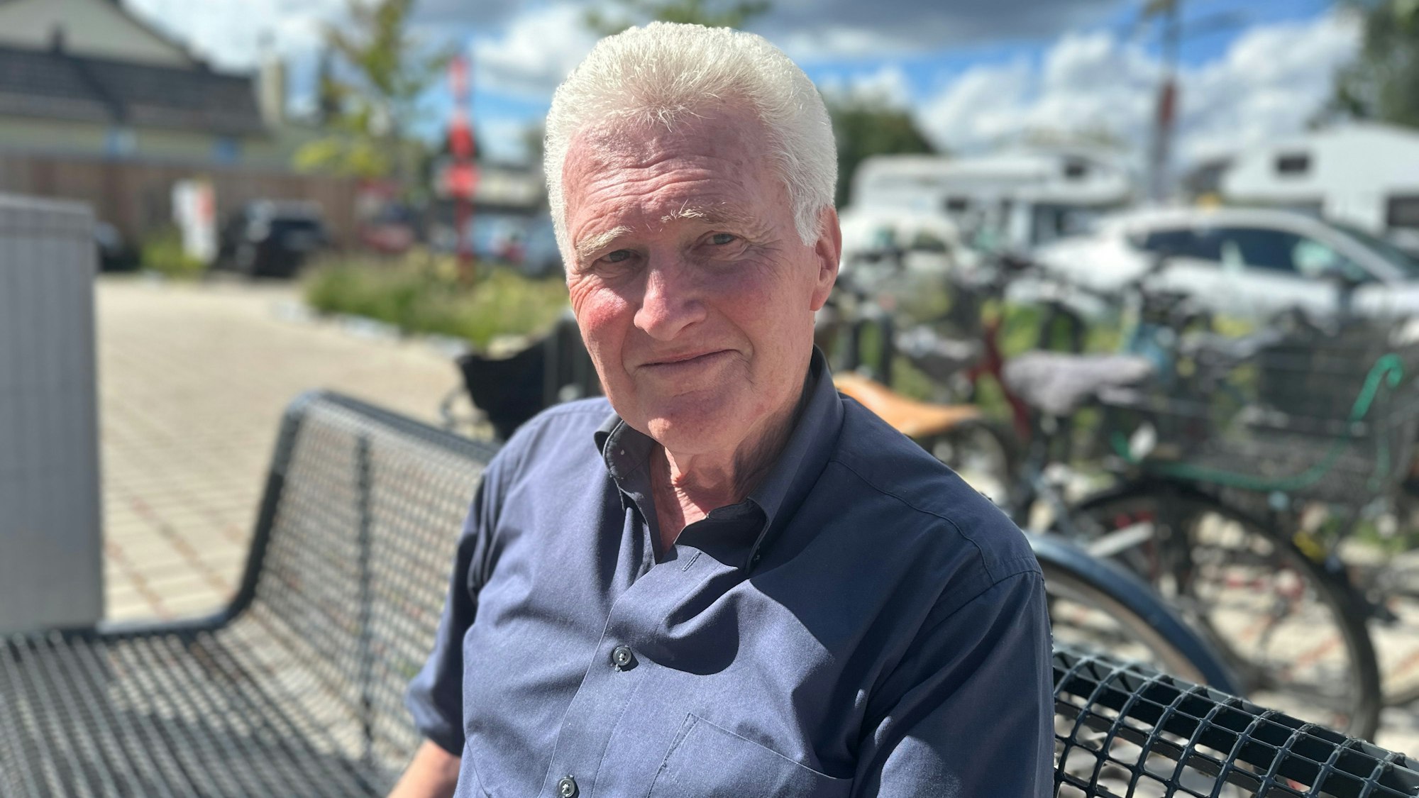 Ein älterer Mann mit weißem Haar trägt ein dunkelblaues Hemd und sitzt auf einer Bank.