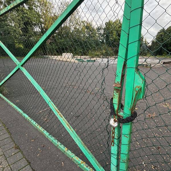 Auf dem Bild ist ein Zaun mit einem Kettenschloss vor einem brachliegenden Grundstück zu sehen.