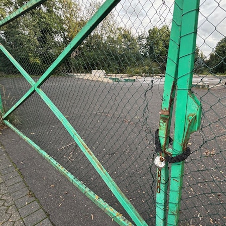 Auf dem Bild ist ein Zaun mit einem Kettenschloss vor einem brachliegenden Grundstück zu sehen.